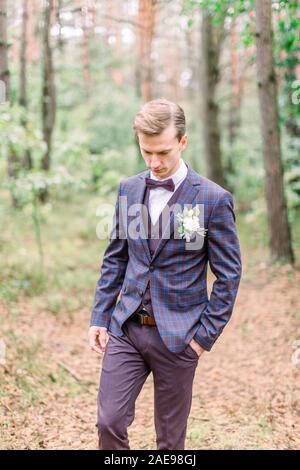 Handsome groom in stylish suit standing on the background of pine forest. Beautiful man, groom posing and preparing for rustic wedding ceremony outdoo Stock Photo