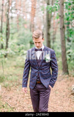 Handsome groom in stylish suit standing on the background of pine forest. Beautiful man, groom posing and preparing for rustic wedding ceremony outdoo Stock Photo
