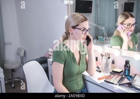 Blond female make up artist talks by phone with client. Professional stylist in her beauty visage studio Stock Photo
