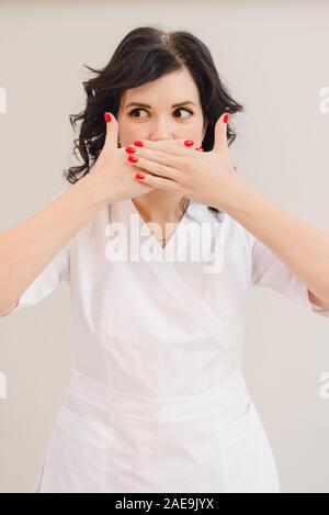 A young girl in a medical coat shows with both hands a sign of silence. A mature brunette model in a cosmetology white suit covered her mouth with her hands on a beige background in the studio. Stock Photo