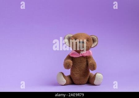 Teddy bear sitting on purple background. Teddy bear toy with pink bow. Copy space, postcard. Plush toy handmade. Stock Photo
