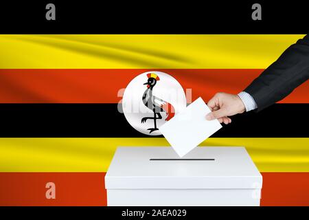 Election in Uganda. The hand of man putting his vote in the ballot box. Waved Uganda flag on background. Stock Photo