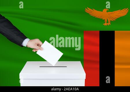 Election in Zambia. The hand of man putting his vote in the ballot box. Waved Zambia flag on background. Stock Photo