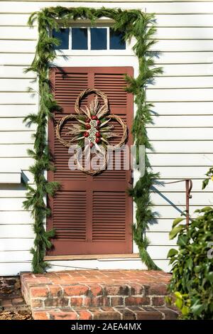 Christmas wreath door decoration, Colonial Williamsburg. Dried Lotus pods, red pomegranates, twisted sticks, on brown louvered door, evergreen swag. Stock Photo