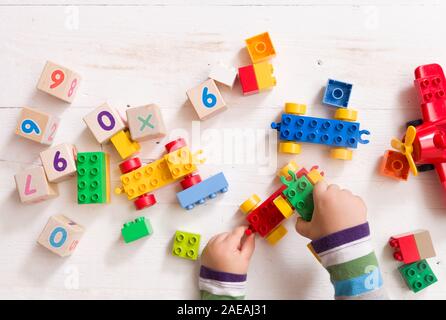 Child playing with wooden cubes with numbers and colorful toy bricks on a white wooden background. Toddler learning numbers. Hand of a child taking to Stock Photo