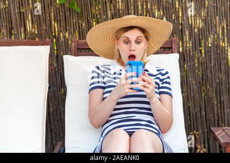 Portrait of suprised young adult blogger woman in hat and dress is sitting on sunbed and holding phone, looking at screen reading news with shocked fa Stock Photo