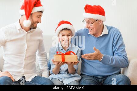 Boy Receiving Christmas Gift From Dad And Grandpa Sitting Indoor Stock Photo