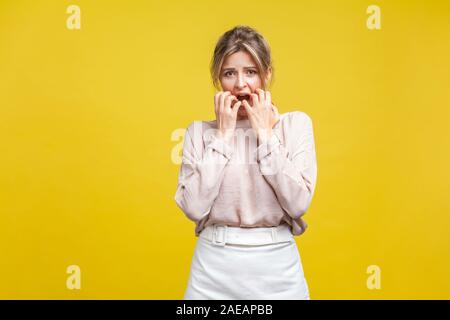 Portrait of nervous, shocked or scared young woman in casual beige blouse standing, looking at camera with hands in mouth and biting nails. indoor stu Stock Photo
