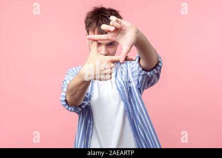 Frame of fingers. Portrait of young man with brunette hair in casual striped shirt looking at camera through hand frame taking picture, focusing the e Stock Photo