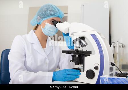 Young laboratory assistant in mask looking through modern microscope Stock Photo