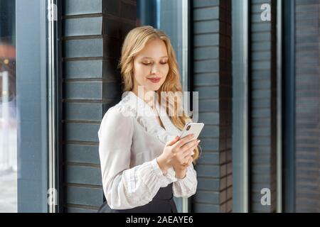 Portrait of stunning elegant businesswoman with long blond hair in white stylish shirt leaning on brick wall and smiling happily while using smartphon Stock Photo