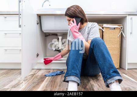 Woman sitting near leaking sink calling for help Stock Photo
