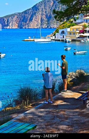 A young couple stand in the shade of an old tree, looking out over the beautiful harbour of the pretty fishing village of Assos. Stock Photo