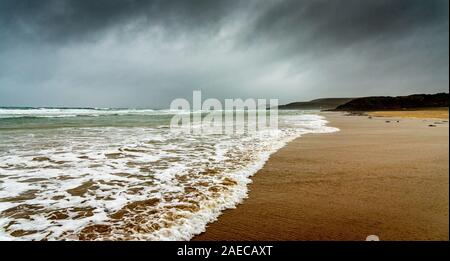 Sandy beach in winter in Brittany. The white foam of the waves of the ocean comes to die on the sand under a dark sky laden with rain. Stock Photo