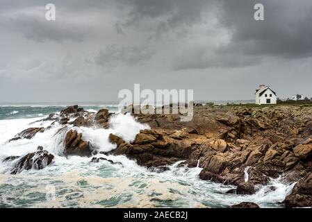 Storm on the coast in Brittany. The waves are smashing on the rocks under a rainy sky. A small white house stands in the wind. Stock Photo