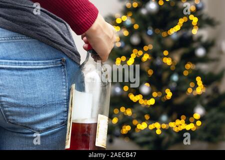 Person in front of the Christmas tree with a bottle of alcohol, drinking too much during Christmas time and New Year, Stock Photo