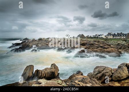 Long exposure on a storm on the coast in Brittany. The foam of the waves crashing on the rocks creates a white fog. The houses of the village face the Stock Photo