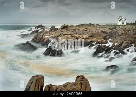 Long exposure on a storm on the coast in Brittany. The foam of the waves crashing on the rocks creates a white fog. The houses of the village stand in Stock Photo