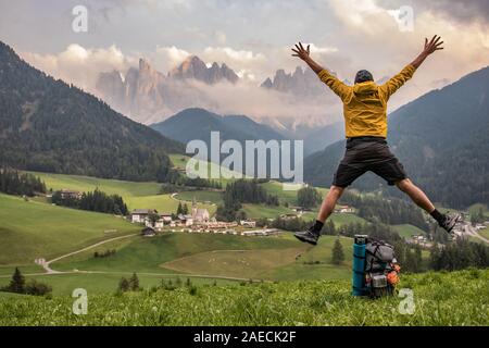 Young man jumping in front of Santa Maddalena. Yellow jacket, backpack, beanie. Dolomites,South Tyrol, Italy Stock Photo