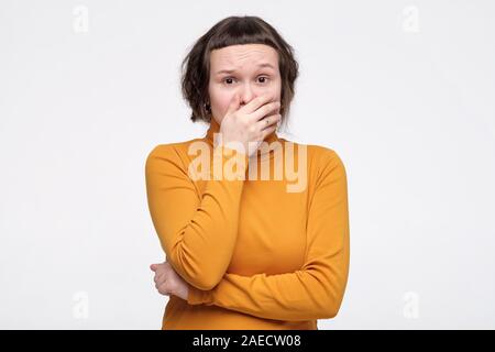 Good looking stunnedteenager in yellow clothes covers mouth with hand stares at camera, tries to be speechless. Studio shot. Stock Photo