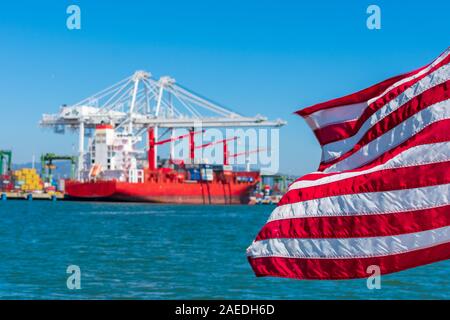 Flag of the United States flying waving beautifully in the wind under blue sky. Blurred container cargo cranes unloading container ship in background Stock Photo