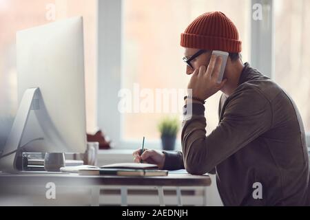 Side view portrait of contemporary businessman wearing glasses and beanie speaking by smartphone while working at desk in office against window, copy space Stock Photo