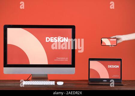 Web design concept with hand holding smartphone, computer and laptop at wooden table over coral background, copy space Stock Photo