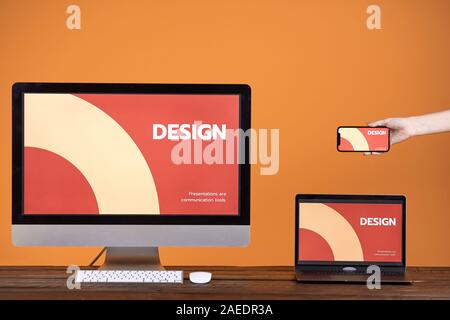 Web design concept with hand holding smartphone, computer and laptop at wooden table over pop orange background, copy space Stock Photo