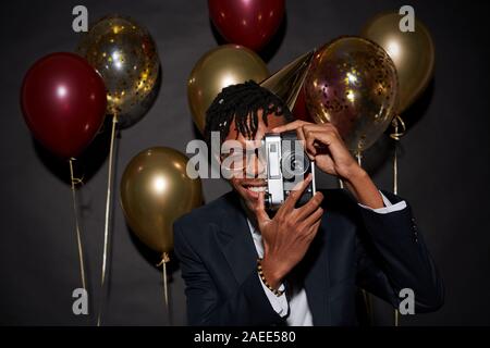 Waist up portrait of contemporary African man holding photo camera while posing against black background with party balloons, shot with flash Stock Photo