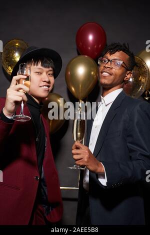 Portrait of two ethnic young men raising glasses to camera while posing against black background at party, shot with flash Stock Photo