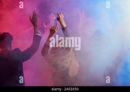 Back view of beautiful young woman dancing and raising hands while enjoying party in smoky nightclub, copy space