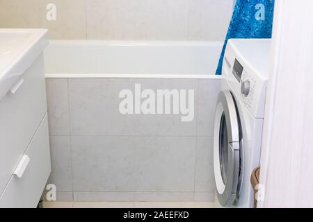 Narrow bathroom in a small apartment decorated with white marble tiles. Covered with a pedestal, a cast-iron bathtub, a washing machine. Small-sized h Stock Photo