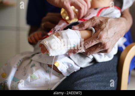 saline infusion. sick baby inward patient sitting on parent legs. baby in hospital being fed. sick kid in treatment process, in hospital and caring by Stock Photo