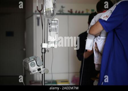 saline infusion. sick baby inward patient sitting on parent legs. baby in hospital being fed. sick kid in treatment process, in hospital and caring by Stock Photo