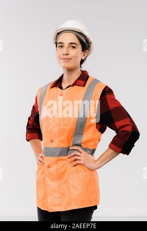 Pretty young female engineer in helmet and orange jacket keeping arms on waist Stock Photo