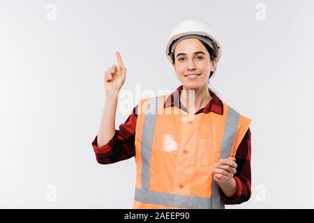 Pretty young female engineer in helmet and orange jacket pointing upwards Stock Photo