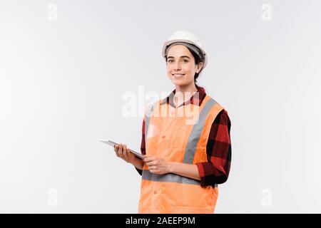 Young cheerful female engineer in helmet and orange jacket using touchpad Stock Photo