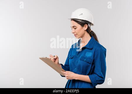Young successful female engineer in blue workwear and safety helmet making notes Stock Photo