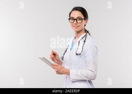 Young female clinician in whitecoat and eyeglasses using touchpad in isolation Stock Photo