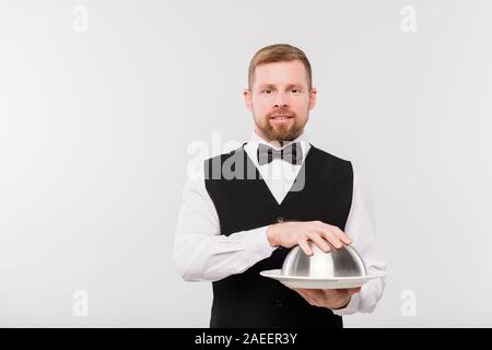 Elegant young waiter in bowtie and black waistcoat holding cloche with food Stock Photo