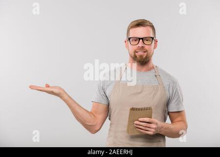 Happy young bearded waiter in apron and eyeglasses showing aside Stock Photo