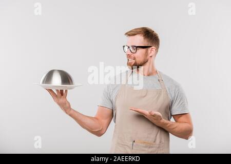 Happy young bearded waiter of cafe in apron and eyeglasses showing cloche Stock Photo