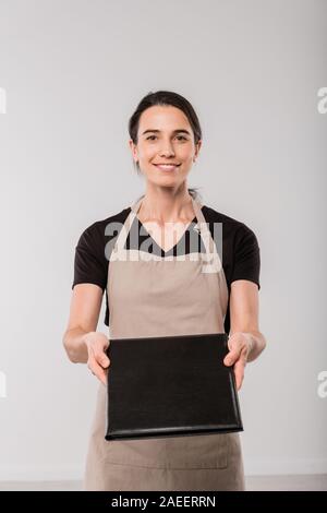 Happy young waitress of cafe in apron giving you menu in black leather cover Stock Photo