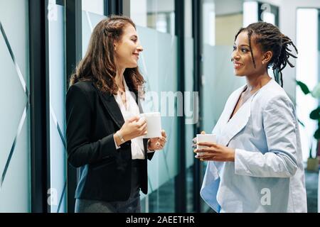 Smiling pretty multi-ethnic business women drinking coffee during break and discussing news and projects Stock Photo