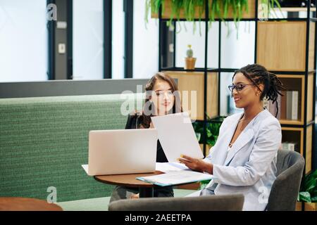 Smiling pretty business lady in glases showing document with financial report to young colleague at meeting Stock Photo