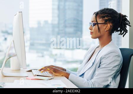 Young stylish Hispanic businesswoman with dreadlocks wearing glasses when working on laptop in office Stock Photo