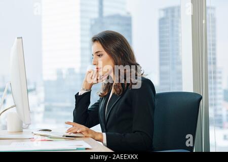 Beautiful serious pensive young businesswoman reading report on computer screen and making conclusions Stock Photo