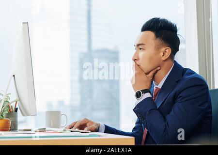 Pensive Vietnamese businessman covering mouth when reading document with important information on computer screen Stock Photo