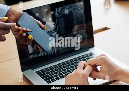 Hands of female project manager showing application on smartphone screen to financial manager working on laptop and making sales report Stock Photo