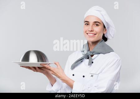 Young smiling female chef in uniform looking at you while carrying cloche Stock Photo